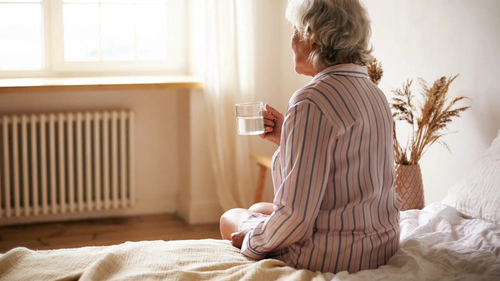 Femme aînée assise sur son lit avec une tasse d’eau dans une chambre lumineuse
