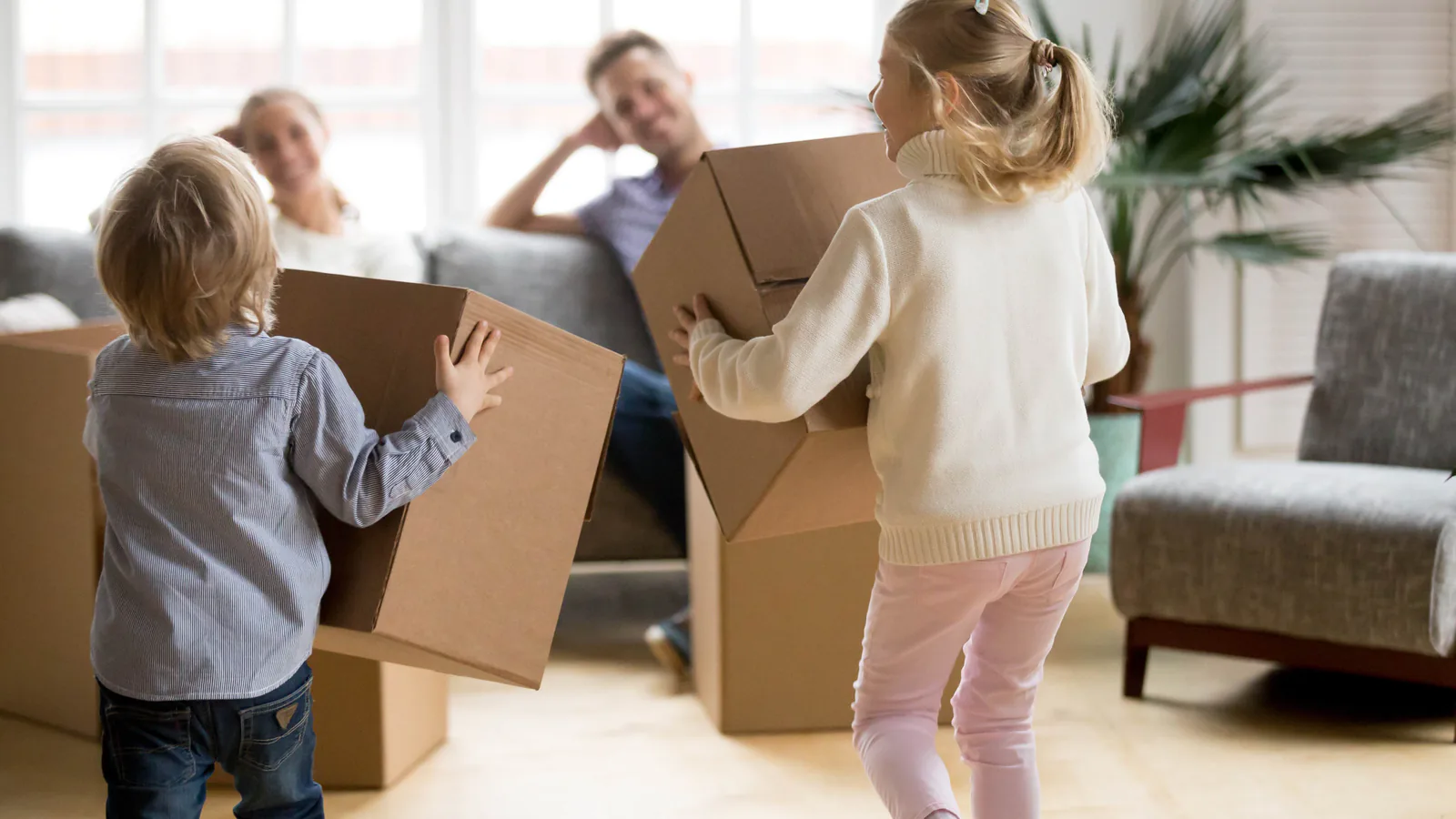 Two children carrying cardboard boxes while their parents sit nearby during a family move