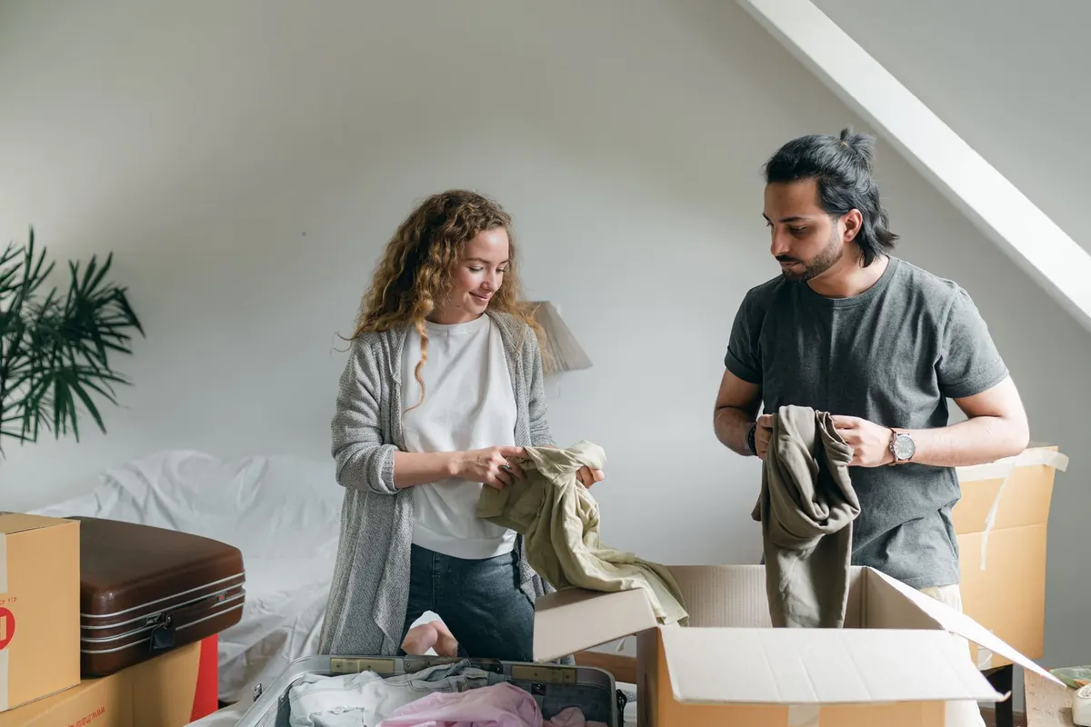 Couple folding clothes beside moving boxes and an open suitcase